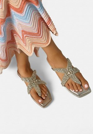 Pies con esmalte de uñas rosa claro que llevan sandalias plateadas en forma de estrella de mar con joyas y el dobladillo visible de un vestido con patrón ondulado multicolor.