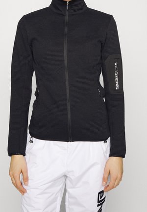 Chaqueta negra de forro polar con textura acanalada vertical, cremallera completa, cuello alto y bolsillos laterales. Presenta un pequeño detalle en la manga.