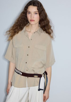 Junge Frau mit langen lockigen Haaren, die ein beiges kurzärmeliges Hemd mit Knöpfen, weiße Hose und einen dunklen Gürtel mit einem gebundenen gemusterten Schal trägt.