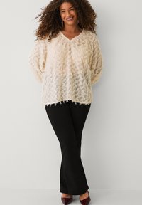 Glunderende vrouw met krullend haar, gekleed in een crèmekleurige, textuurachtige V-hals trui, zwarte pantalons en bordeauxrode hakken, staand tegen een effen lichte achtergrond.