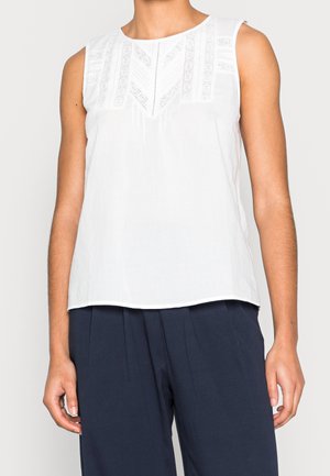 Blouse blanche sans manches avec panneaux en dentelle sur le plastron avant, portée avec un pantalon plissé bleu marine.