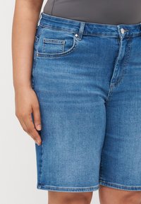 Modri jeans kratke hlače s srednjim pasom, sprednjimi žepi in ravnim krojem. Tkanina ima gladko teksturo in minimalno zbledelost.