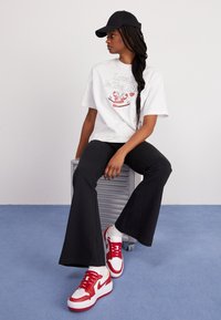 Bílá tričko s grafickým potiskem, černé rozšířené kalhoty a červeno-bílé tenisky. Černá čepice, sedící na stříbrném vozíku na světlém pozadí.