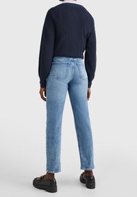 Persona vista di spalle con un maglione blu navy, jeans dritti azzurri e mocassini neri con dettaglio in catena dorata, in piedi su uno sfondo chiaro.