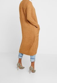 En person som bär en lång, beige cardigan med fickor, blå cropped jeans och högklackade skor med orm-mönster, står mot en enkel vit bakgrund.