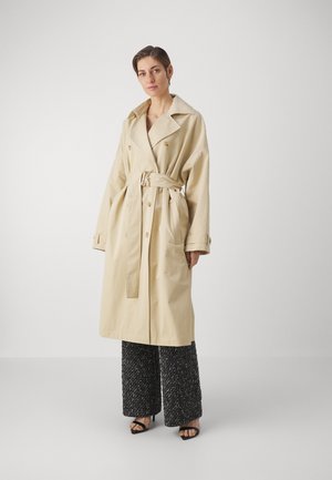 Light beige trench coat with a large collar, belted waist, double-breasted buttons, and side pockets. Model wears textured black wide-legged pants.
