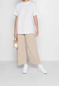 Bílá bavlněná trička s krátkým rukávem a centrálním logem, v kombinaci s béžovými širokými kalhotami a bílými teniskami. Malá kabelka s hnědými akcenty.
