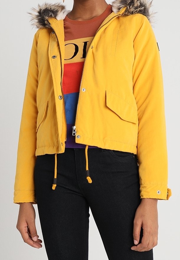 Femme portant une veste jaune à capuche bordée de fourrure, par-dessus une chemise imprimée multicolore et un pantalon noir, debout devant un fond uni.