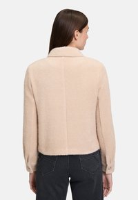 Kobieta widziana z tyłu, ubrana w beżową, strukturalną kurtkę z długimi rękawami oraz ciemne, wysokie dżinsy.