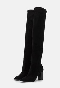 Botas de ante negras por encima de la rodilla con punta afilada y tacones gruesos, con una textura suave y detalles mínimos.