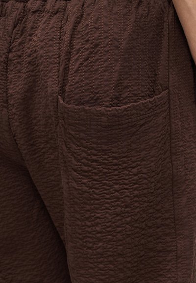 Κοντινή λήψη υφασμάτινου παντελονιού σκούρου καφέ με ανάγλυφη υφή, που δείχνει μια πλαϊνή τσέπη και μέρος του χεριού ενός ατόμου που ακουμπάει στη μέση.