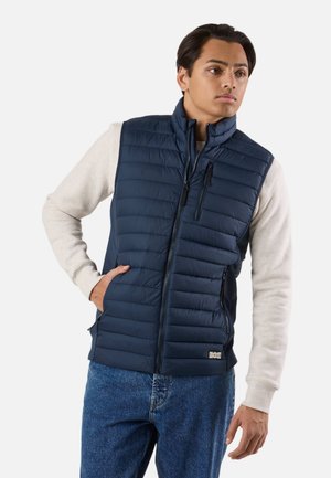 Young man wearing a navy padded vest over a light beige long-sleeve shirt and blue jeans, standing with one hand in vest pocket.