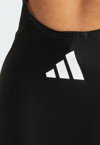 Crni sportski top s glatkom teksturom, s bijelim Adidas logotipom koji se sastoji od tri dijagonalne trake.