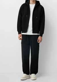 Muž v černé zipové mikině s kapsami, bílém tričku, černých cargo kalhotách a bílých teniskách, stojící na jednolitém bílém pozadí.