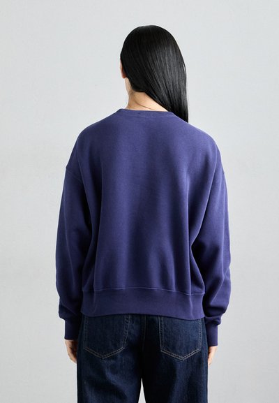 Sudadera morada de corte relajado, con puños y dobladillo de canalé, que presenta un clásico cuello redondo, combinada con jeans de mezclilla oscura.