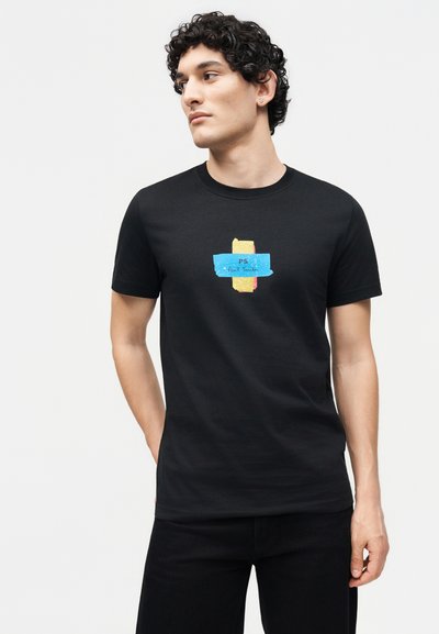 Jeune homme aux cheveux bouclés portant un t-shirt noir avec un motif abstrait bleu et jaune et le texte « PS Paul Smith ».
