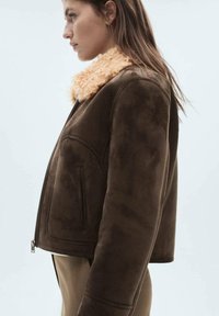 Jachetă din suede maro cu un guler pufoș bej, având fermoare laterale și un design ajustat. Textură netedă, formă structurată.