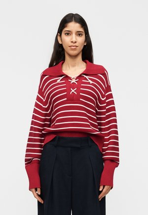 Donna che indossa un maglione a righe rosse e bianche con colletto stringato e polsini oversize, abbinato a pantaloni neri a vita alta, in piedi.