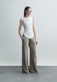 Femme portant un haut blanc sans manches, un pantalon beige à jambes larges, des lunettes de soleil, debout les mains dans les poches devant un fond uni.