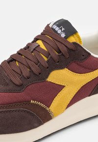 Sneaker in suede e tessuto marrone con accenti bordeaux, gialli e beige, dotata di lacci robusti e di un logo a contrasto sulla linguetta.