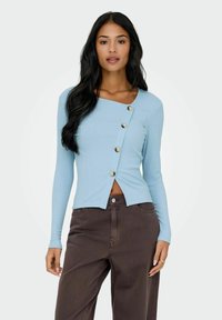 Jasnoniebieska, prążkowana bluzka z długim rękawem o asymetrycznym dole i dekoracyjnymi guzikami z przodu. Połączona z ciemnobrązowymi, szerokimi spodniami.