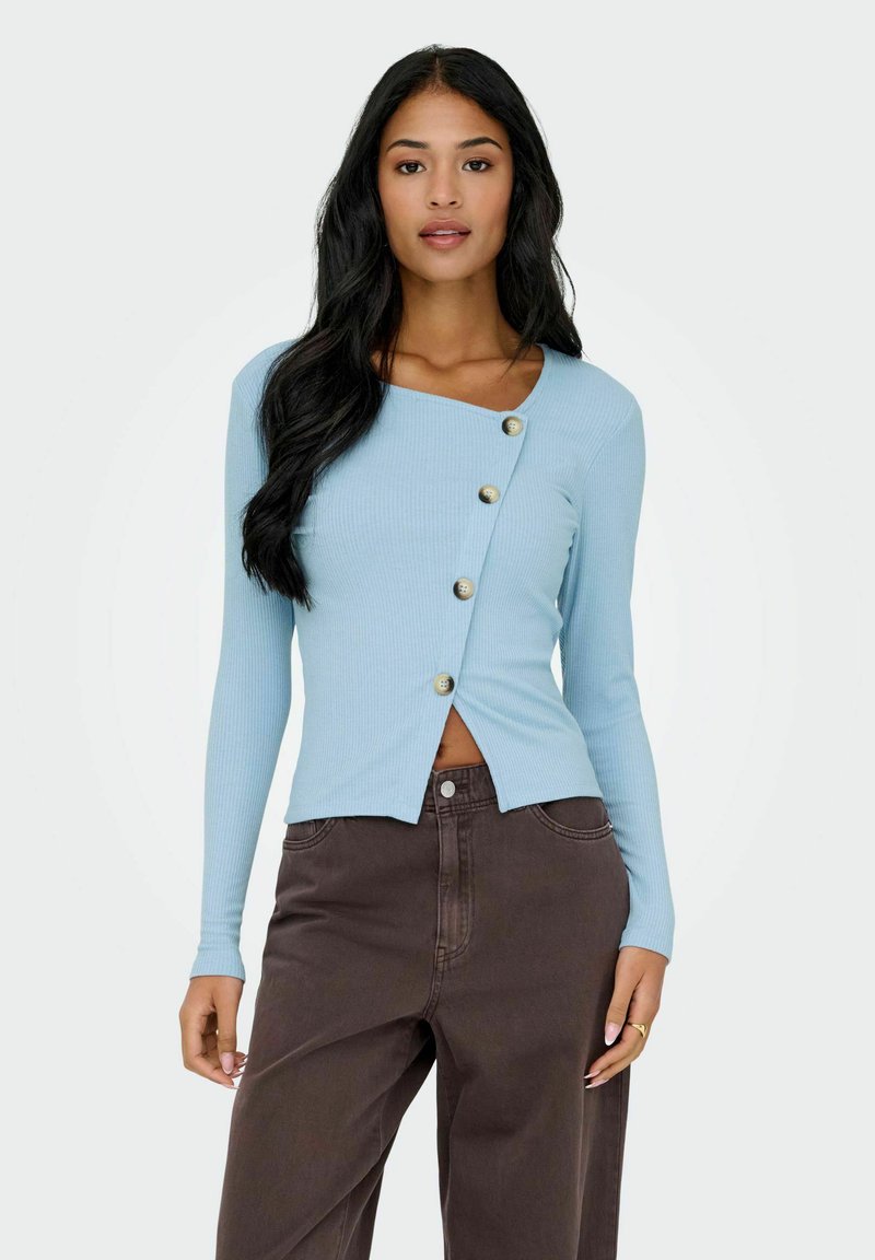 Jasnoniebieska, prążkowana bluzka z długim rękawem o asymetrycznym dole i dekoracyjnymi guzikami z przodu. Połączona z ciemnobrązowymi, szerokimi spodniami.