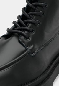 Černá kožená bota s kulatou špičkou, texturovanými tkaničkami a kontrastním modrým prošíváním podél okrajů. Hladký, lesklý povrch s dírkami pro tkaničky.