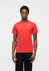 Άνδρας που φοράει απλό κόκκινο κοντομάνικο μπλουζάκι με μικρό μπλε και λευκό λογότυπο στο αριστερό στήθος, σε  συνδυασμό με μαύρο παντελόνι.