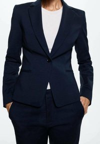 Blazer azul marino de tejido texturizado, con un solo botón, solapas normales y bolsillos laterales minimalistas. Combinado con una blusa blanca.