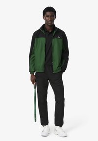 Giacca a zip verde e nera, polo nera, pantaloni neri, sneakers bianche. In mano una racchetta da tennis verde. Design atletico casual.