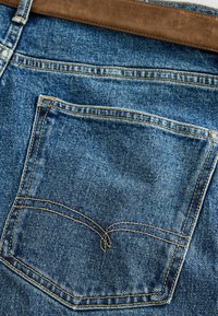 Poche arrière d'un jean en denim bleu avec une double couture en fil orange et une ceinture en daim marron à la taille.