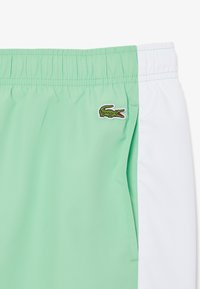 Shorts de bain vert menthe avec un panneau latéral blanc, une taille élastique et un logo de crocodile vert brodé sur le devant à gauche.