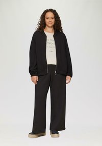 Schwarze Zip-Jacke aus strukturiertem Material, getragen über einem hellen Hemd mit grünem Text. Kombiniert mit lockeren schwarzen Hosen und Plateauschuhen.