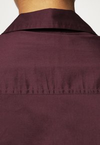 Camisa burdeos con cuello clásico y una textura sutil, con detalles fruncidos en la parte posterior para un interés de diseño adicional.