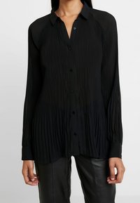Blusa nera a maniche lunghe con pieghe, chiusura frontale con bottoni e collo con risvolto. Presenta una texture morbida e un orlo curvo, abbinata a pantaloni in pelle nera.