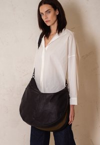 Borsa a tracolla nera in nylon con fondo curvo, tracolla regolabile e design a strati; esposta da un modello che indossa una maglietta bianca.