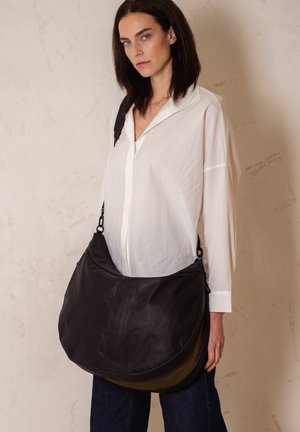 Borsa a tracolla nera in nylon con fondo curvo, tracolla regolabile e design a strati; esposta da un modello che indossa una maglietta bianca.