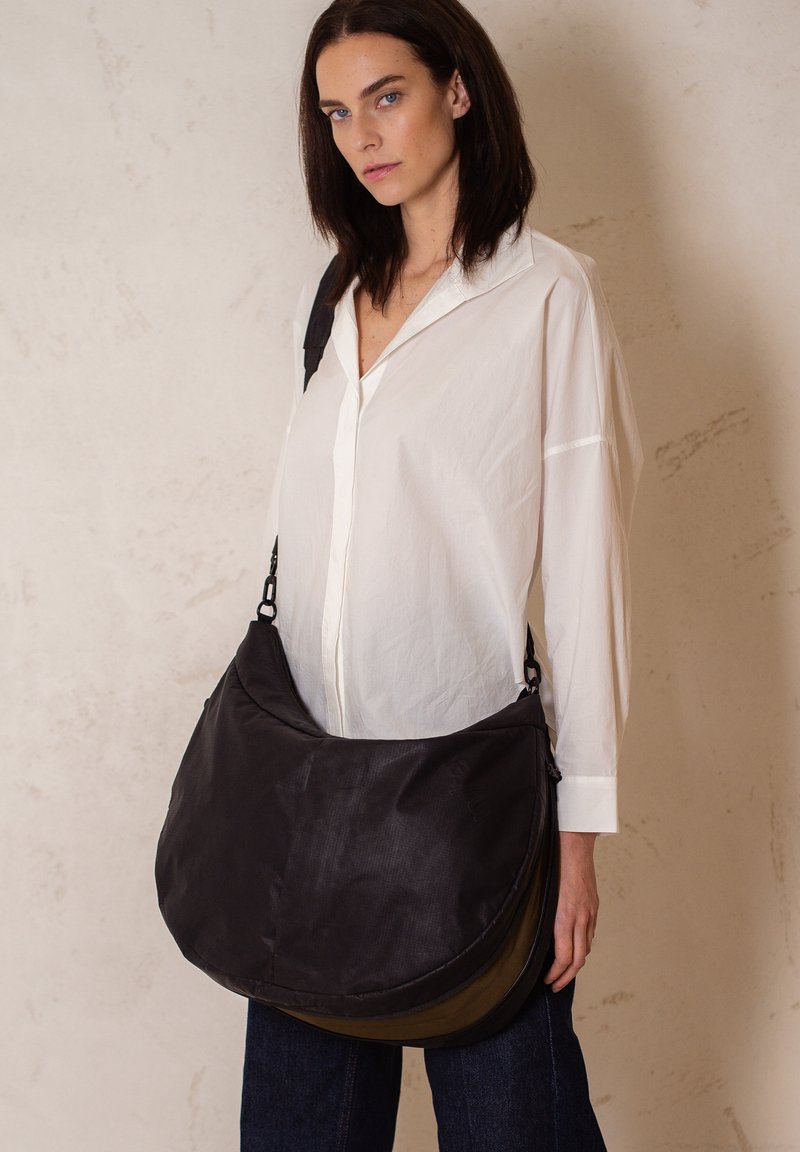 Borsa a tracolla nera in nylon con fondo curvo, tracolla regolabile e design a strati; esposta da un modello che indossa una maglietta bianca.