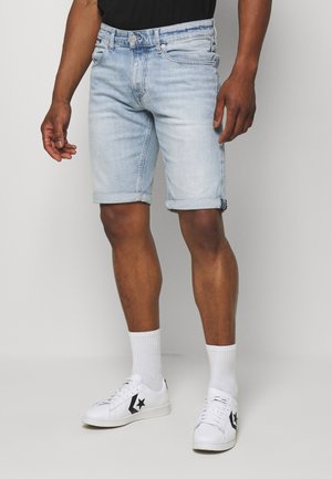 Homme portant un short en jean bleu clair roulé, chaussettes blanches et baskets blanches avec un logo étoile noir, debout devant un fond uni.