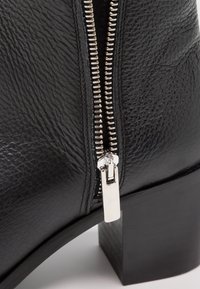 Stivale alla caviglia in pelle nera con cerniera argentata, finitura texturizzata e tacco spesso resistente. La vista ravvicinata mette in evidenza il dettaglio della cerniera.