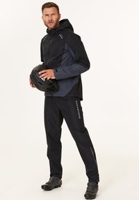 Waterdichte jas in zwart en grijs, met een capuchon en ritsen. Zwarte broek met reflecterende accenten. Model houdt een zwarte helm vast.