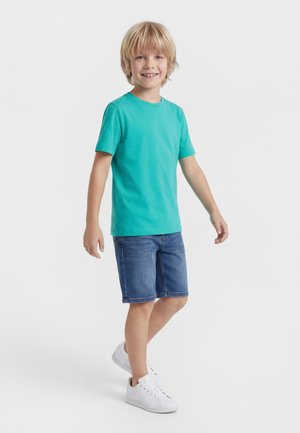 Χαμογελαστό ξανθό αγόρι που φοράει τιρκουάζ μπλουζάκι, τζιν σορτς και λευκά αθλητικά παπούτσια, στέκεται με ένα πόδι ελαφρώς σηκωμένο πάνω σε απλό λευκό φόντο.