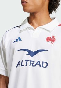 Hvid polo skjorte med ribbet krave, blå detaljer, et kyllingeemblem og "ALTRAD" i mørkeblåt på tværs af brystet.