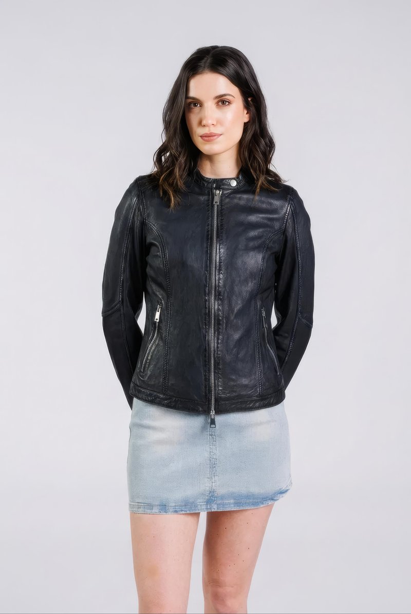 Vrouw met donker golvend haar, gekleed in een zwart leren jasje met ritssluiting en een lichtblauwe denim minirok, staand tegen een effen lichte achtergrond.