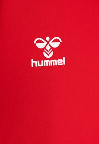 T-shirt de sport rouge au texture lisse avec un logo Hummel blanc proéminent, représentant des ailes stylisées et une forme semblable à un arbre.