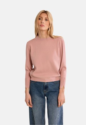 Donna che indossa un maglione rosa a maniche lunghe con bottoni sui polsini e jeans blu morbidi, in piedi davanti a uno sfondo chiaro e semplice.