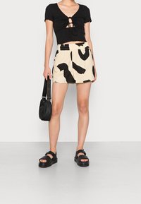 Black short-sleeve top with a cinched front, paired with cream shorts featuring a bold black abstract pattern. Black handbag and sandals.