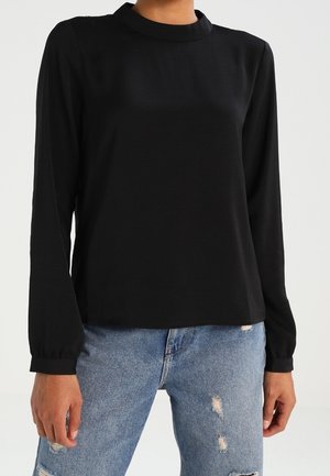 Maglia nera a maniche lunghe con scollatura a girocollo e texture liscia, abbinata a jeans blu strappati. Maniche prominenti con polsini arricciati.