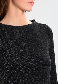 Černý svetr s texturovaným povrchem a metalickými akcenty, s kulatým výstřihem a dlouhými rukávy, zdůrazňující přiléhavý design.