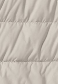 Polsterstoff in hellem Beige, mit glatter Textur und subtil glänzender Oberfläche, sowie gesteppten Abschnitten, die ein puffiges Erscheinungsbild erzeugen.